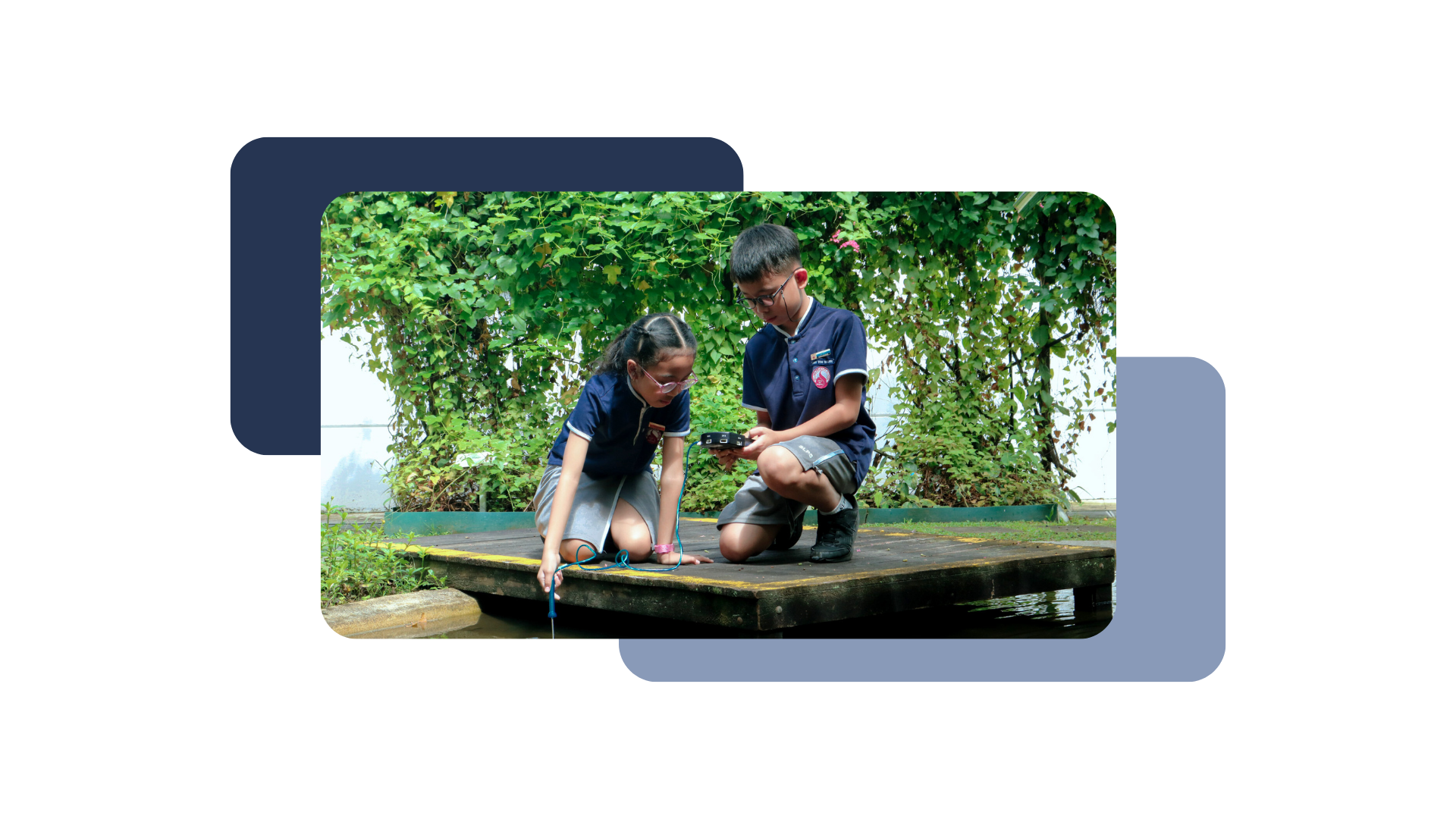 Two students in school uniforms kneeling on a wooden platform by a pond, conducting a science experiment outdoors by lowering a sensor into the water while one student holds a measuring device.